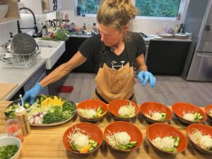 A chef wearing an apron and blue gloves prepares vibrant rice bowls in a modern kitchen, carefully arranging fresh vegetables from a large platter into individual red bowls.