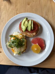 Breakfast plate with a toasted sandwich topped with avocado and tomato on one half, fried egg, cheese, and arugula on the other half, served with slices of grapefruit and orange on a white plate