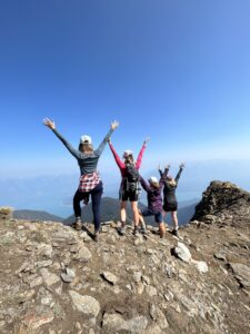 Four women celebrate at the summit of Sentry Peak in the Esplanade Range, British Columbia, with panoramic views of alpine valleys and lakes below.