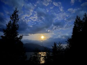 Full moon rising over a lake in the Kootenays in British Columbia, framed by silhouetted trees and dramatic evening clouds.