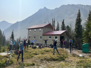 A group of people doing outdoor yoga in front of Meadow Hut, nestled among alpine trees and rugged mountains in the Esplanade Range, British Columbia.