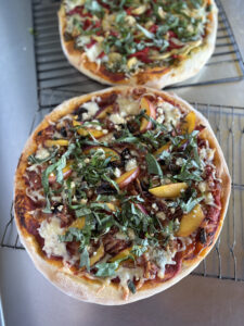 Two freshly baked gourmet pizzas on cooling racks, topped with vibrant ingredients including sliced peaches, basil, blue cheese, mushrooms, and assorted vegetables.