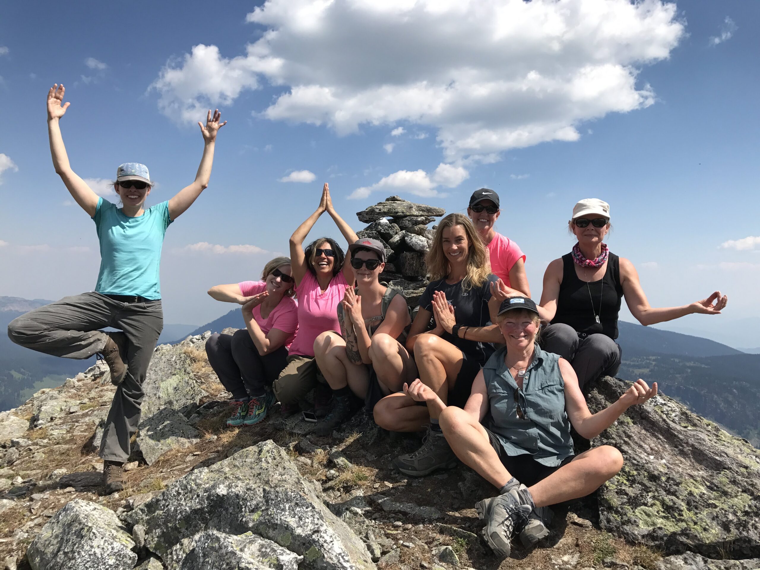 eight women posing  on top of a mountain. One woman is holding tree pose.