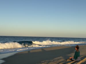 Woman sitting on the beach with waves crashing in the background. The woman is meditating looking out at the Pacific Ocean