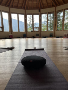 A yoga mat with a meditation cushion is positioned in a spacious circular studio with wooden floors and large windows offering views of forest and mountain peaks at Kootenay Lake, Kaslo, B.C.