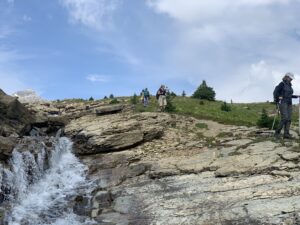 Hikers descend a rocky alpine trail beside a cascading waterfall at Mistaya Lodge near Golden, B.C., surrounded by lush greenery and mountain views under a blue sky.