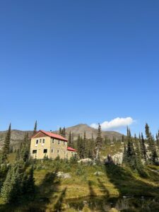 A beige backcountry lodge with a red roof sits in an alpine meadow surrounded by evergreen trees, with rugged mountain peaks and a clear blue sky in the background.