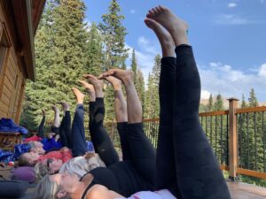 A group of women lying on yoga mats with their legs extended vertically in the air on a wooden deck, surrounded by tall pine trees and mountains under a clear blue sky at Mistaya Lodge.