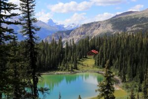 A red-roofed mountain lodge nestled in dense forest beside a turquoise alpine lake, with rugged mountain peaks and glaciers in the background under a partly cloudy sky.