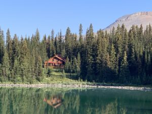 A wooden backcountry lodge nestled in dense evergreen forest, reflected in a still alpine lake under a clear blue sky. A rocky mountain peak rises in the background.