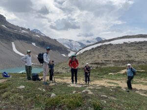 Group of hikers pause on an alpine trail overlooking a glacial lake and snow-capped peaks near Mistaya Lodge, British Columbia, during a summer hiking retreat with Revelstoke Wellness