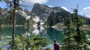 A hiker in a pink shirt and sunglasses smiles at the camera while standing in front of a turquoise alpine lake, surrounded by evergreen trees and snow-capped mountains under a bright blue sky in Mt. Revelstoke National Park