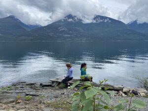 Two women sit on driftwood facing Kootenay Lake, surrounded by misty mountain peaks and calm waters, during a contemplative moment at a wellness retreat in Kaslo, B.C.