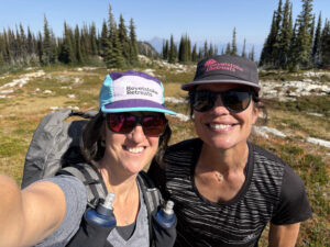 Two women smile for a selfie while hiking in the alpine, both wearing Revelstoke Retreats hats and sunglasses, with evergreen trees and rocky meadows in the background under a clear blue sky.