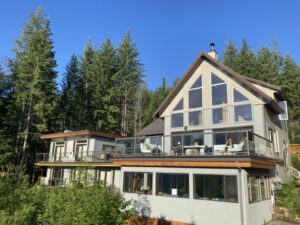 A modern lodge-style retreat centre with large windows and glass railings, nestled in a forested hillside. Two people relax on the upper deck under bright blue skies, surrounded by tall evergreens.