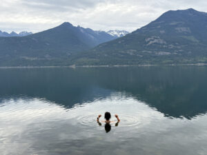 Person enjoying a peaceful swim in calm, glassy water at the Sentinel Retreat Centre in the Kootenays, B.C., surrounded by forested mountains and overcast skies.