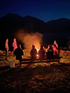 A group of people bundled in warm layers sits and stands around a glowing campfire at night, silhouetted against the dark outlines of distant mountains and a still lake in the Kootenays under a clear starry sky.