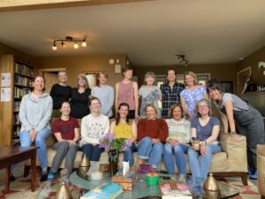A group of 16 women smile together in a cozy living room, gathered around two couches and a coffee table filled with books, candles, and flowers—capturing the warmth and connection of a shared retreat experience.