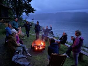 Group of women sitting around a glowing campfire at dusk by the lake during the Sentinel Yoga & Journaling Retreat in the Kootenays, British Columbia.