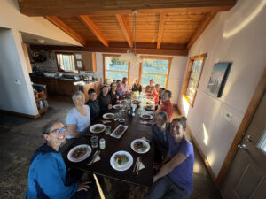 A joyful group of yoga retreat participants seated around a long dinner table in a cozy mountain lodge, sharing a homemade meal at sunset.