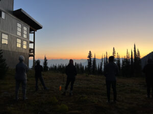 A group of silhouetted people stand outside Sentry Lodge at sunrise, gazing toward a glowing sky and distant mountain peaks framed by evergreens in the B.C. backcountry.