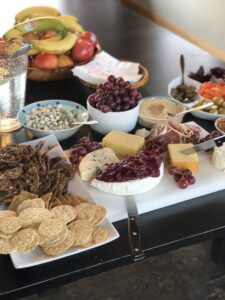 A rustic charcuterie spread featuring a variety of cheeses, crackers, grapes, dried fruit crisps, hummus, olives, pickled vegetables, and fresh fruit, set on a wooden table at a mountain lodge wellness retreat.