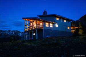 A remote mountain lodge lit warmly from within, surrounded by alpine wilderness and snow-capped peaks at twilight in the Esplanade Range, British Columbia.