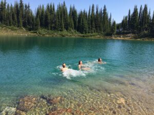 Three women swimming in a crystal-clear alpine lake at Sol Mountain Lodge in the Monashee Range during the 2016 Hiking & Yoga Retreat, surrounded by evergreen forest and bright blue skies.