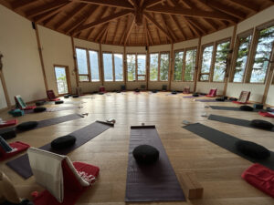 A circular yoga studio with wooden beams and large windows overlooking Kootenay Lake, set up with mats, cushions, and journals for a yoga and journaling retreat in Kaslo, B.C., 2019.
