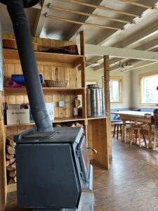 A cozy cabin interior at Vista Lodge featuring a wood-burning stove, stacked firewood, open shelves with yoga mats and a guitar, and a communal dining table with forest views through the windows.