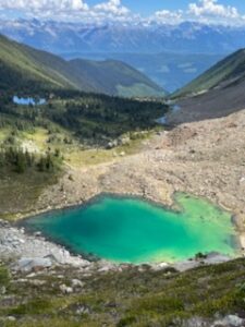 A turquoise alpine lake nestled in a rocky basin, surrounded by green valleys and distant mountains under a blue sky. Vista Lodge is visible in the distance, tucked into the Esplanade Range.