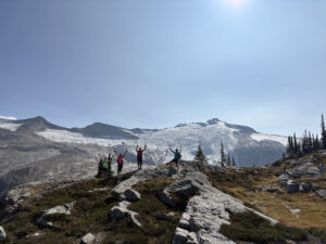 Group of women raising trekking poles in celebration with Blanket Glacier in the background, during the 2025 Local Revelstoke Retreat in the Monashee Mountains west of Revelstoke, BC
