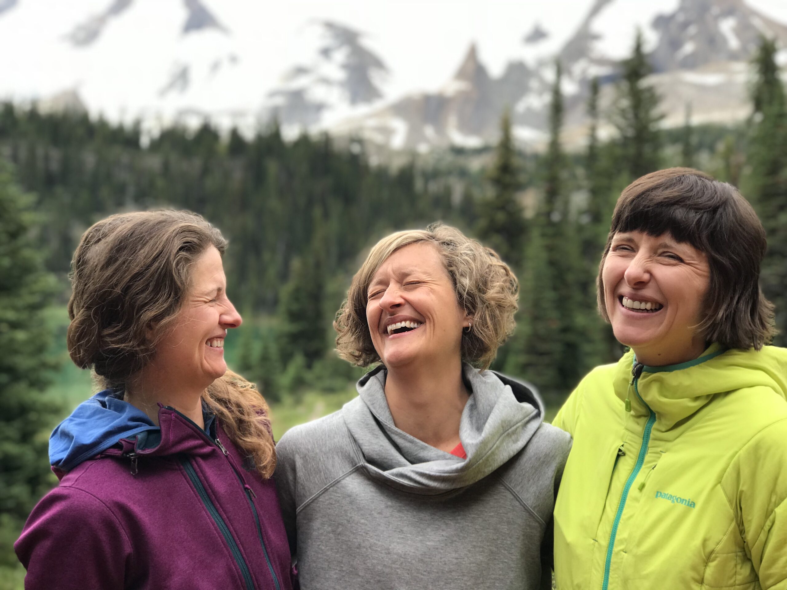 three women in hiking gear smile and laugh. The Canadian Rockies are visible in the backgrounds.