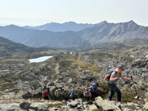 a group of women on a wellness retreat in BC with hiking backpacks and equipment on. There is a scenic rocky mountain backdrop and alpine lake. Photo taken somewhere along Rasta Pass.