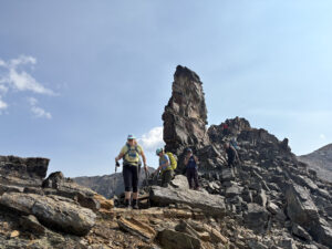 A group of women hiking along a rocky alpine ridge in the Canadian Rockies on a sunny day, with trekking poles and backpacks, during a Revelstoke Retreats adventure above Unicor Lake