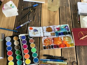 Picture of a few different water color paint sets sitting on a table with journals around.