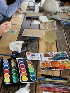 Picture of a few different water color paint sets sitting on a table with journals around.